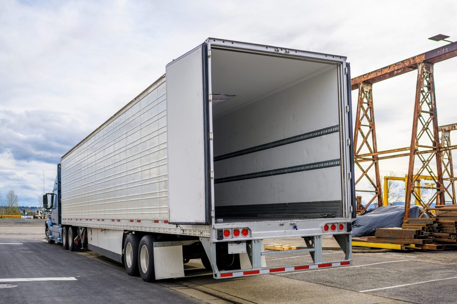 Professional grade Big rig blue semi truck with open door empty semi trailer standing at warehouse parking lot at industrial area waiting for the commercial load for the next delivery